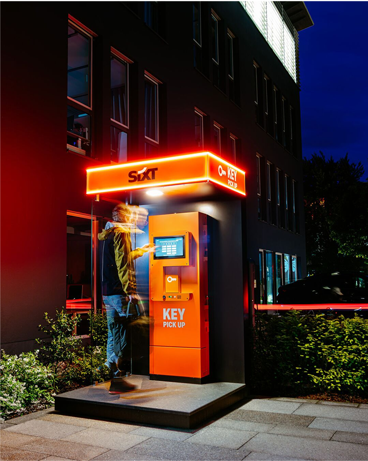 An illuminated self-service key pick-up pillar outside a closed branch at night, with a customer reaching for the screen